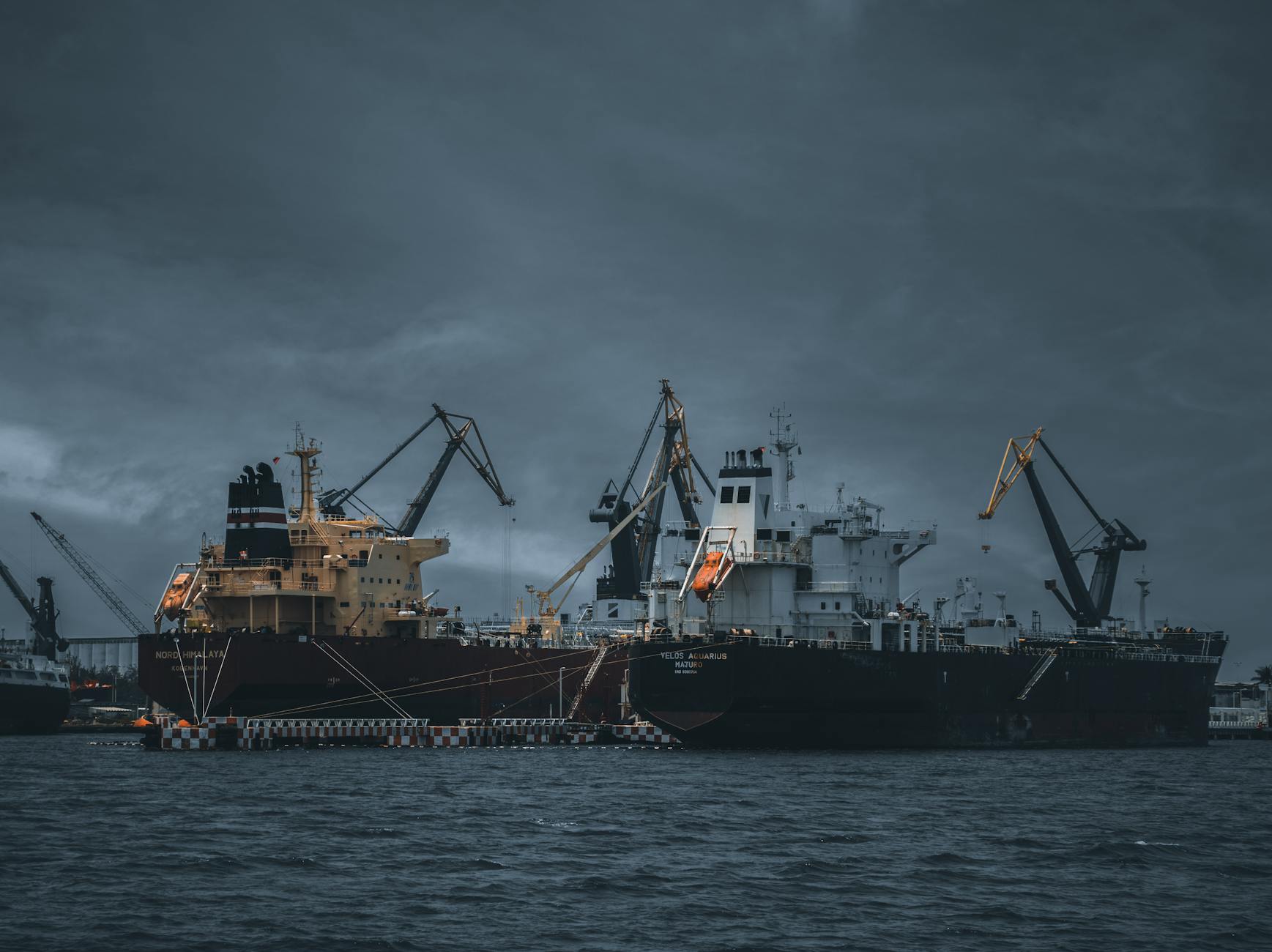 Large cargo ship docked at harbor, cranes and antennas visible under dark cloudy sky.
